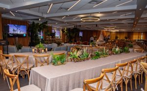 wooden chairs and linen draped tables with greenery as centerpieces fill a banquet room with soft amber lighting