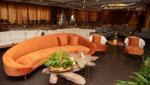Banquet room with amber colored low sofa and chair with decorative oak coffee table in the foreground, with raised seating rows of linen draped tables behind.