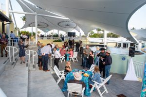 white outdoor chairs surround blue fabric-draped tables under white sun shade panels with a group of people mingling in front of a large outdoor aquarium habitat