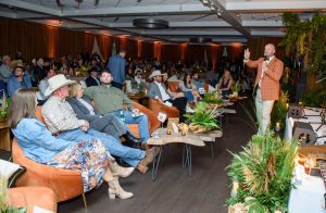A group of men and women in cowboy-inspired attire lounge on rust-colored couches facing a male event host in a rust-colored jacket.